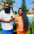 Two men, one with a beard and the other with long hair, holding Hakki-Pikki_Herbal_Hair_Oil bottles outdoors.