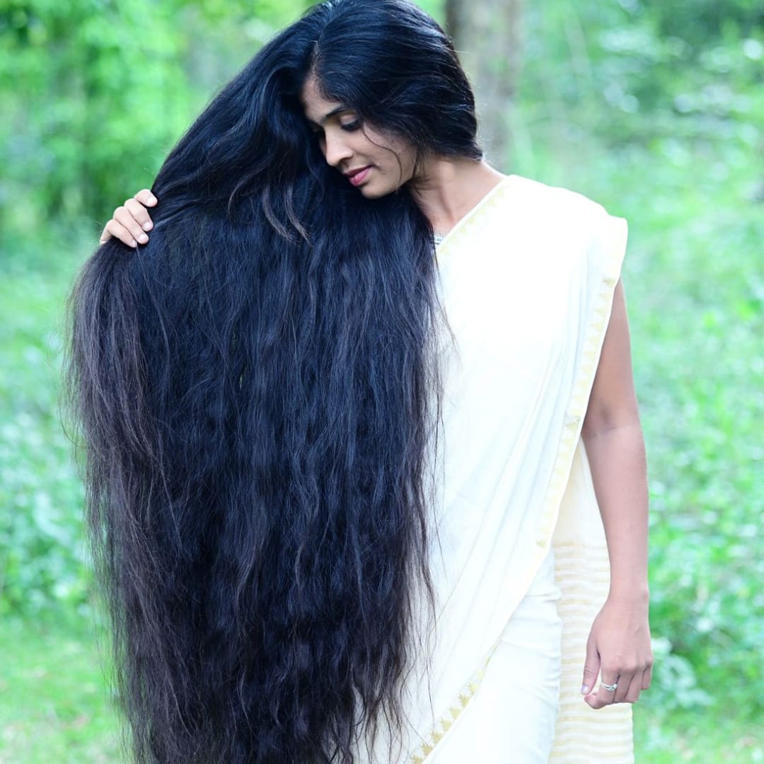 Woman with long black hair wearing a white saree against a green outdoor background