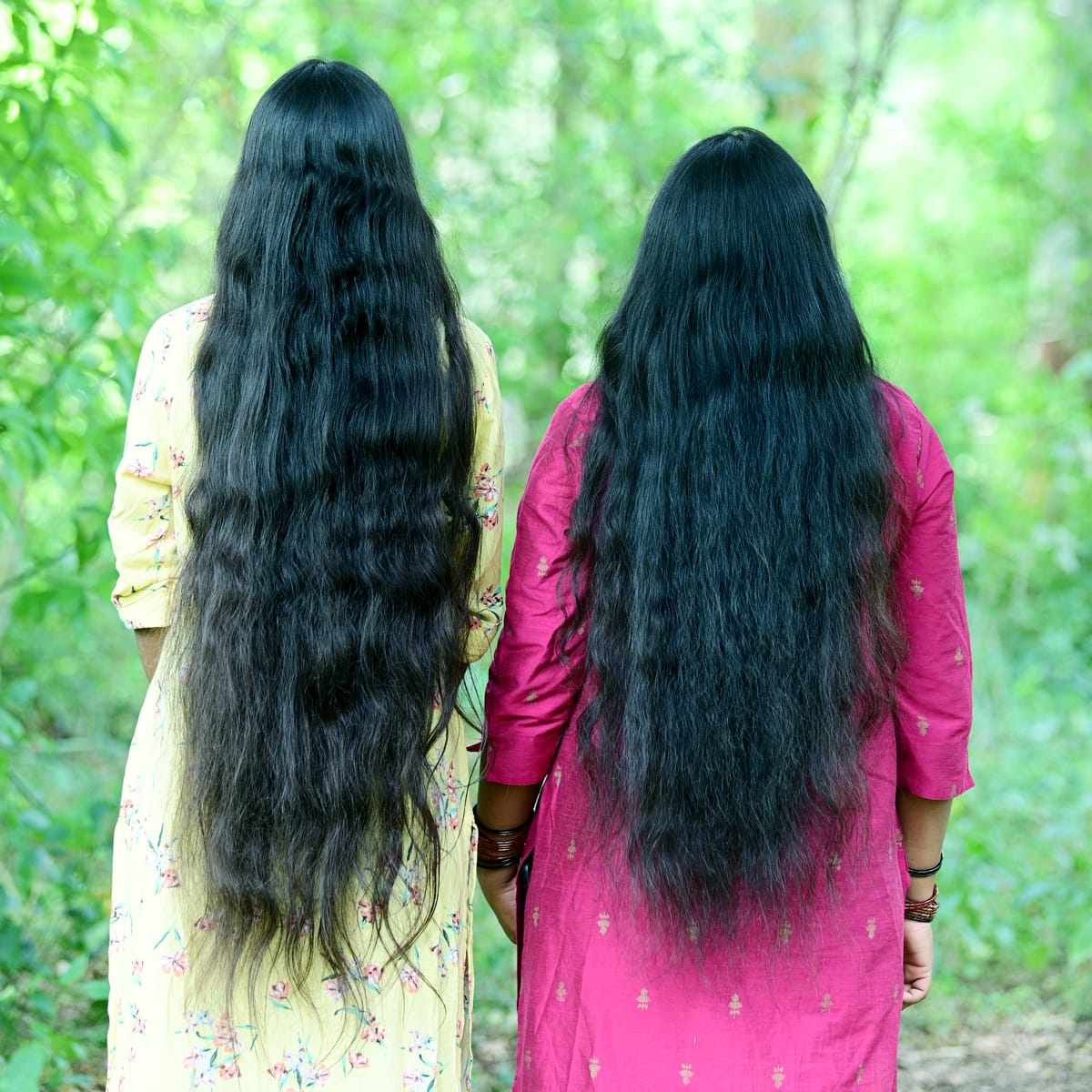 Two women with long, black hair standing back-to-back in a forest setting.