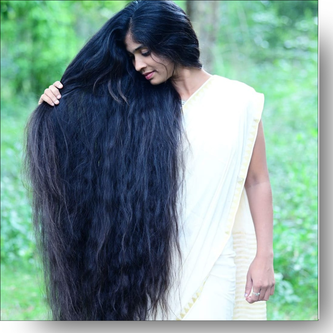 Woman with long black hair wearing a white saree against a green outdoor background